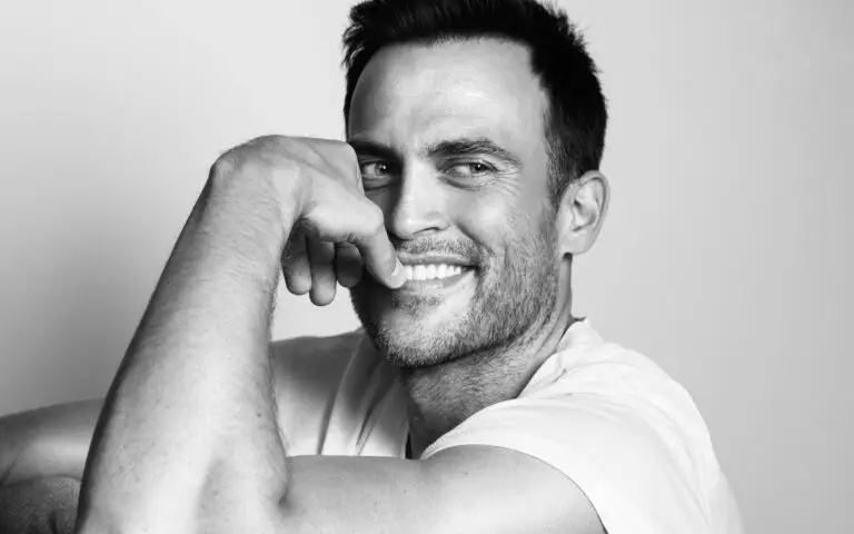 Cheyenne Jackson smiling in a black and white press photo, white t-shirt, arm raised