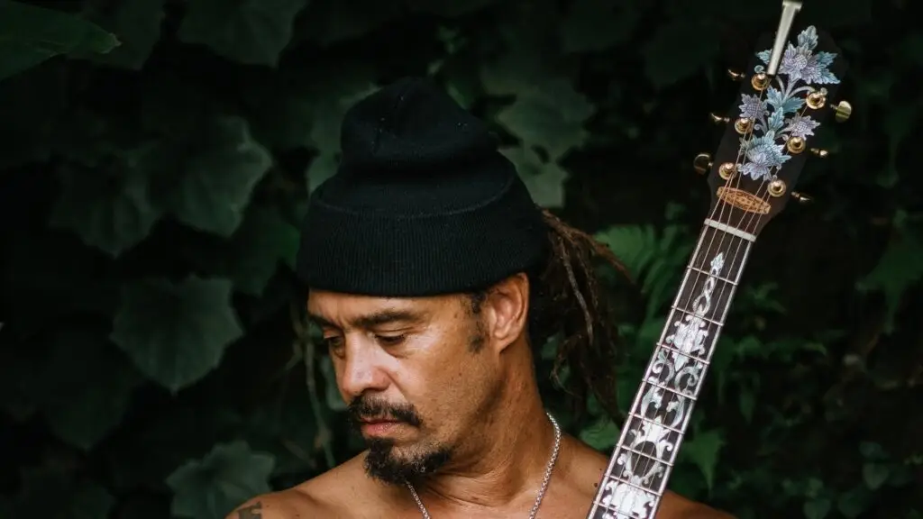 Michael Franti holding an acoustic guitar with lush green foliage background