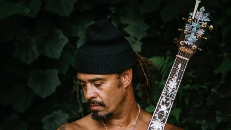 Michael Franti holding an acoustic guitar with lush green foliage background
