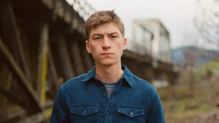 Sam Barber stands before a weathered railroad trestle, denim shirt, country folk press photo