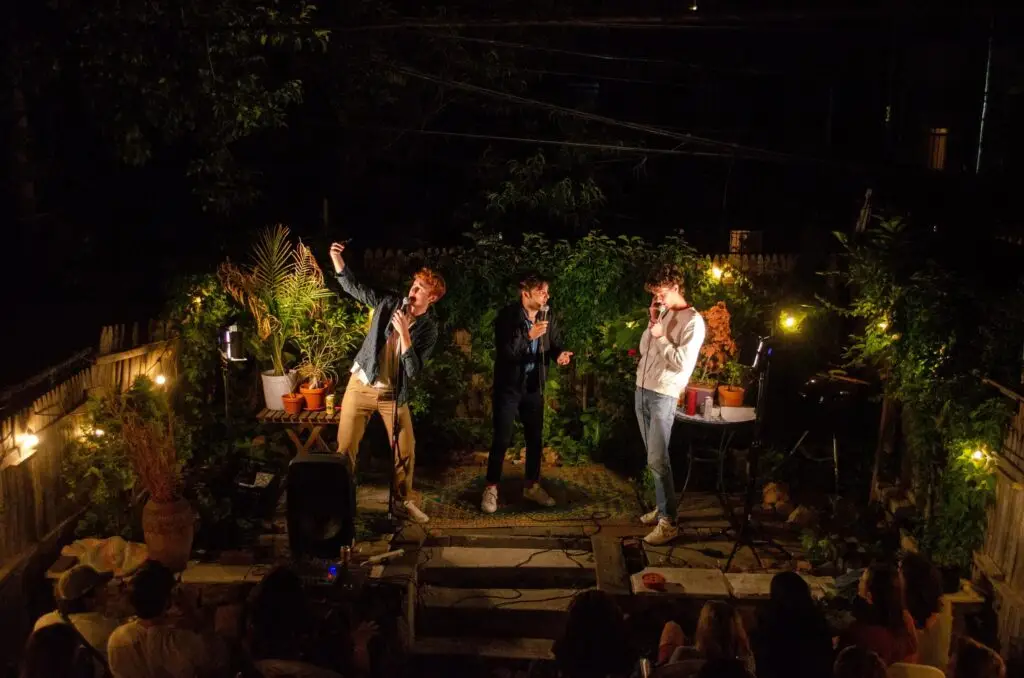 Please Don't Destroy performing live at night on an outdoor garden stage, all three members at microphones