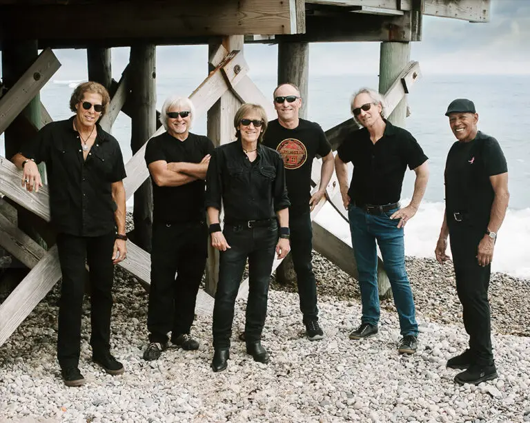 John Cafferty & The Beaver Brown Band on a rocky beach, all six members in black with sunglasses