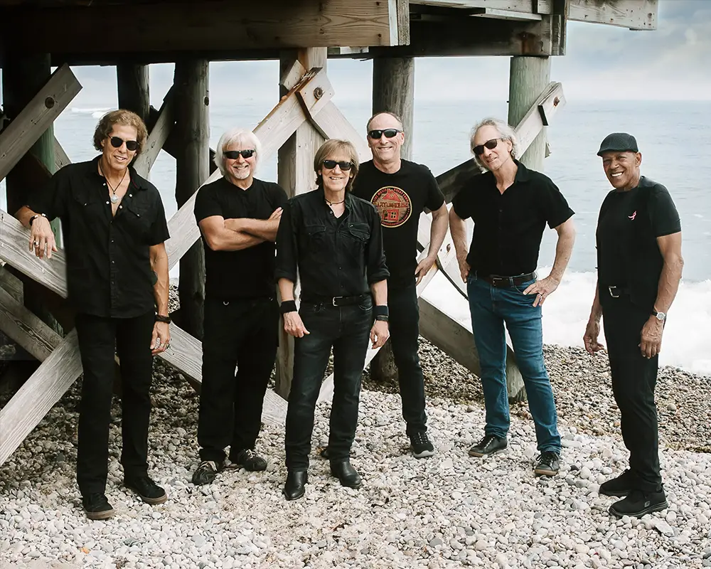 John Cafferty & The Beaver Brown Band on a rocky beach, all six members in black with sunglasses