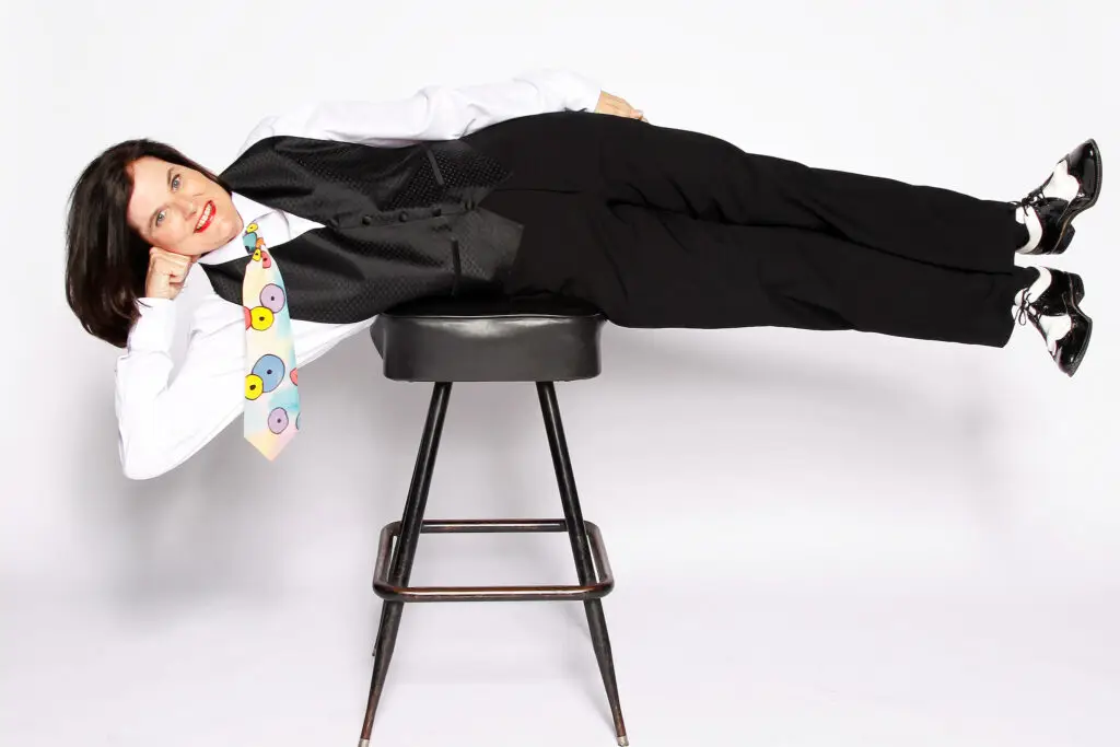 Paula Poundstone comedian balancing horizontally on a bar stool in a tuxedo vest and colorful polka-dot tie