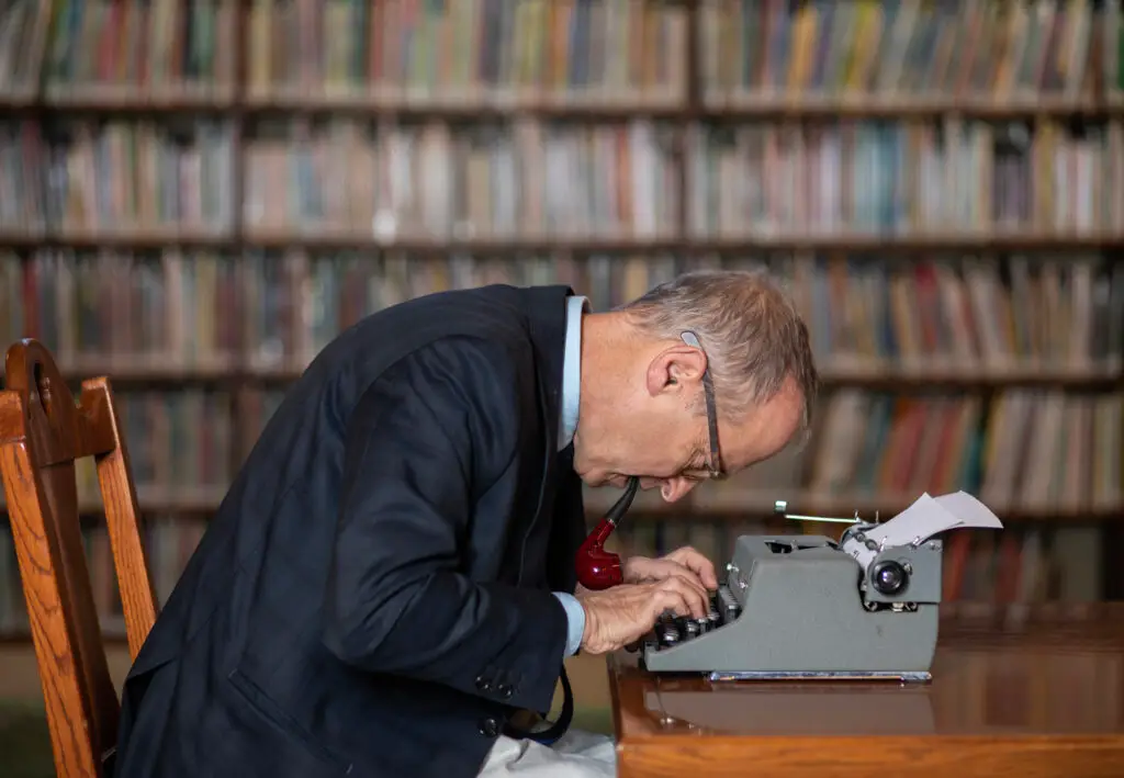 David Sedaris leaning over a typewriter with a pipe, surrounded by floor-to-ceiling library bookshelves