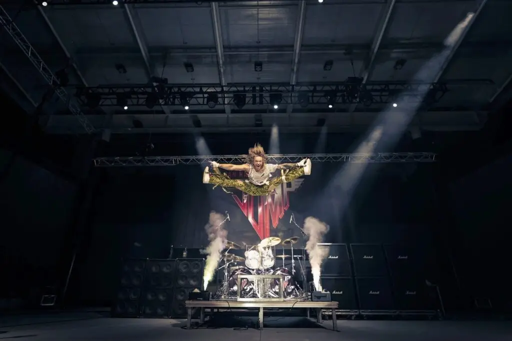 JUMP performer leaping in mid-air split on stage in Van Halen-era costume with Marshall amp stacks and JUMP logo banner backdrop