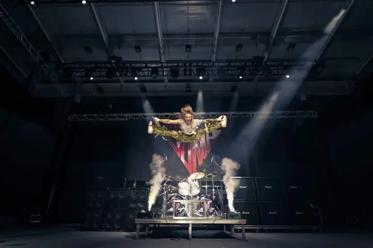 JUMP performer leaping in mid-air split on stage in Van Halen-era costume with Marshall amp stacks and JUMP logo banner backdrop