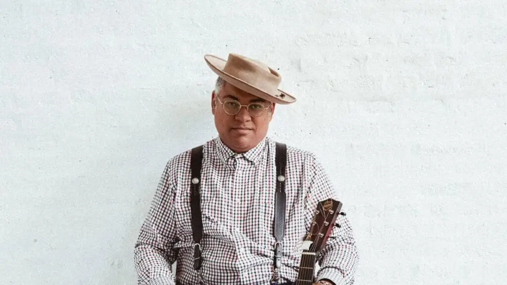 Dom Flemons holding resonator guitar in wide-brim hat and suspenders, official press photo