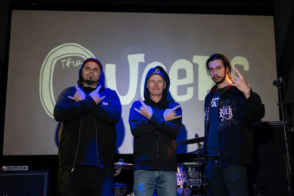The Queers band members posing in front of their logo backdrop at a live venue