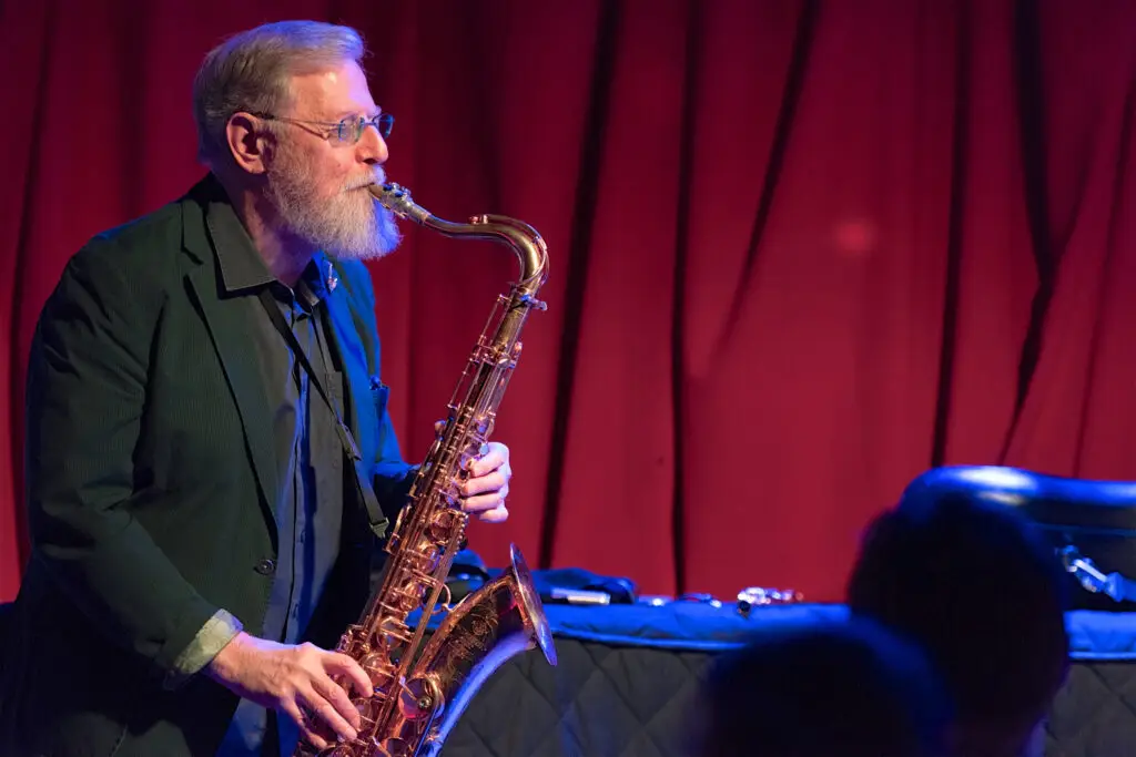 Lew Tabackin playing tenor saxophone on stage, red curtain backdrop, jazz performance