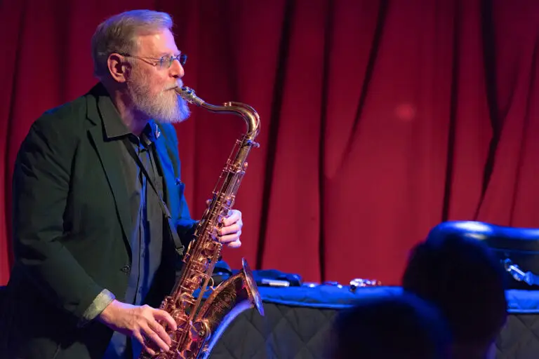 Lew Tabackin playing tenor saxophone on stage, red curtain backdrop, jazz performance