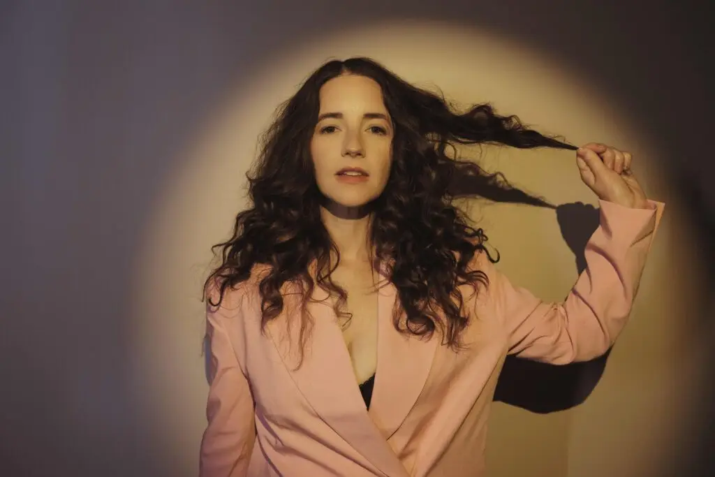 Michaela Anne solo press photo, curly dark hair, pink blazer, studio portrait