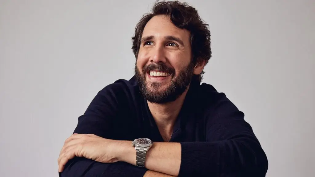 Josh Groban smiling in a studio press photo, dark curly hair and beard, navy hoodie