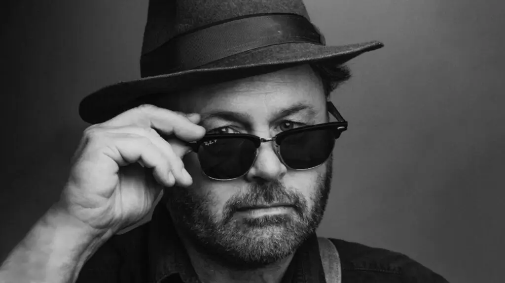 Martin Sexton in a black-and-white press photo, adjusting his Ray-Ban sunglasses beneath a fedora hat