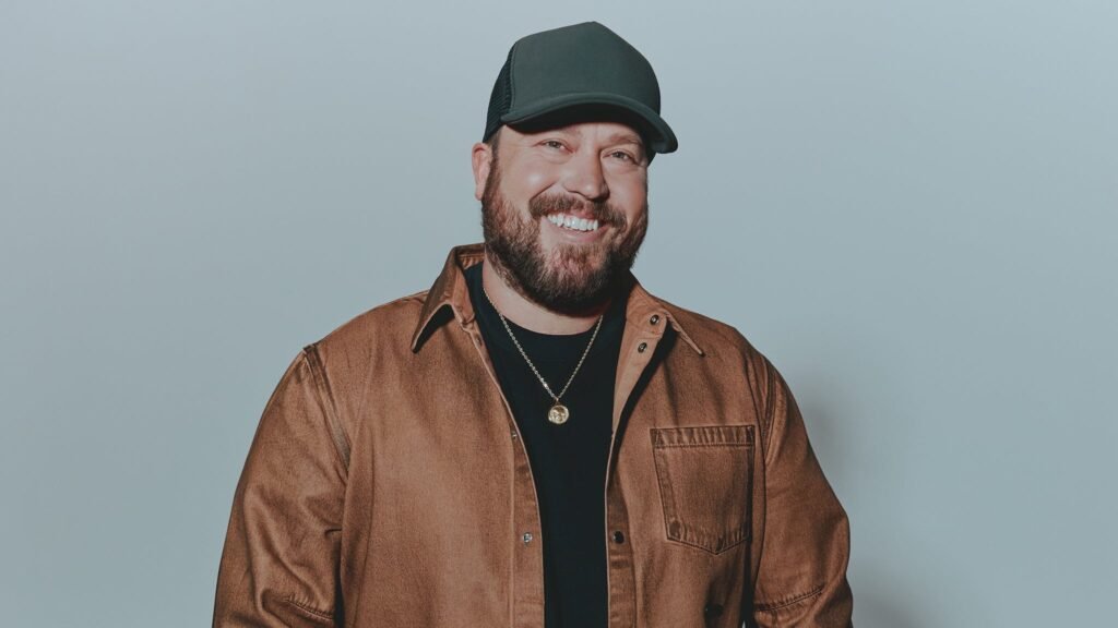 Mitchell Tenpenny in an official press photo, smiling in a green trucker cap and brown jacket