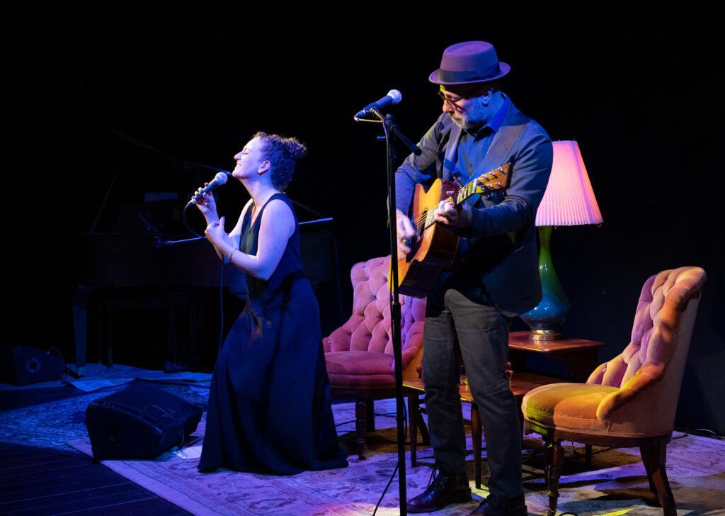 Both Sides Now performers Danielle Wertz and Robbie Schaefer on stage in theatrical cabaret setting with velvet armchairs
