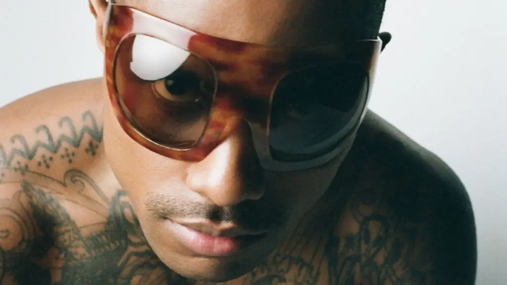 Steve Lacy press photo wearing oversized tortoiseshell sunglasses, tattooed chest visible