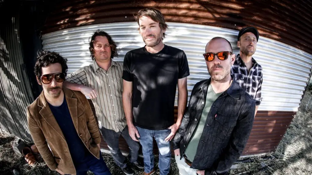 Mad Caddies band promo photo, five members posed outdoors against corrugated metal wall