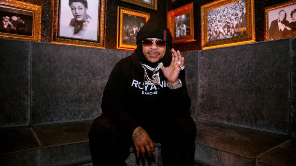 Max B poses in a music venue wearing a black beanie and iced chain necklace