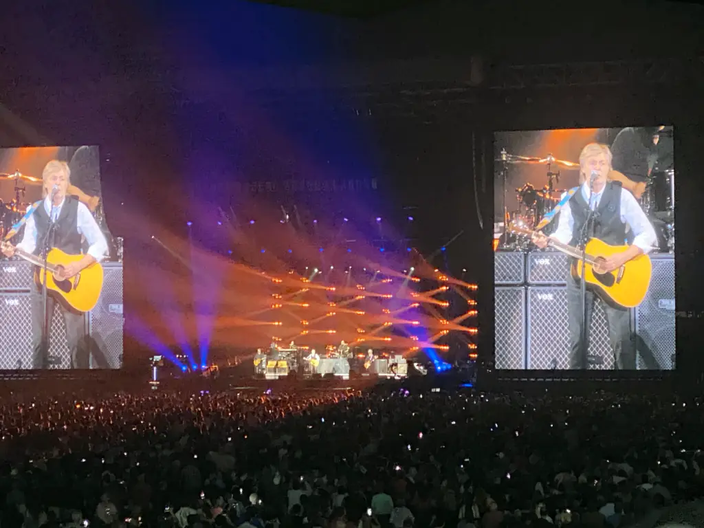 A packed JMA Wireless Dome bowl during a Paul McCartney concert, twin video screens showing McCartney with an acoustic guitar