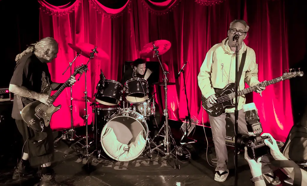 Flipper performing live, guitarist and vocalist on stage with red curtains, intimate punk venue