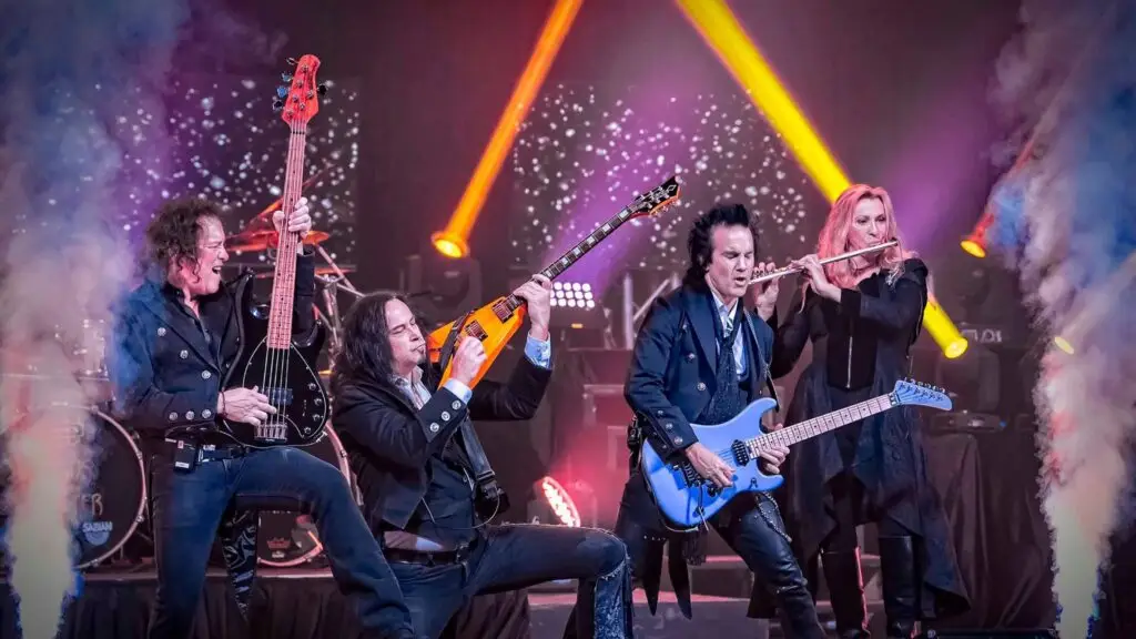 Wizards of Winter performing live — rock ensemble with guitarists and flutist under theatrical stage lighting
