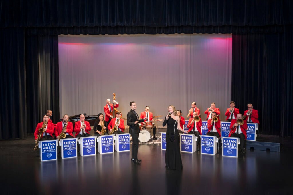 Glenn Miller Orchestra performing on stage, full 15-piece big band in red jackets with vocalists at center