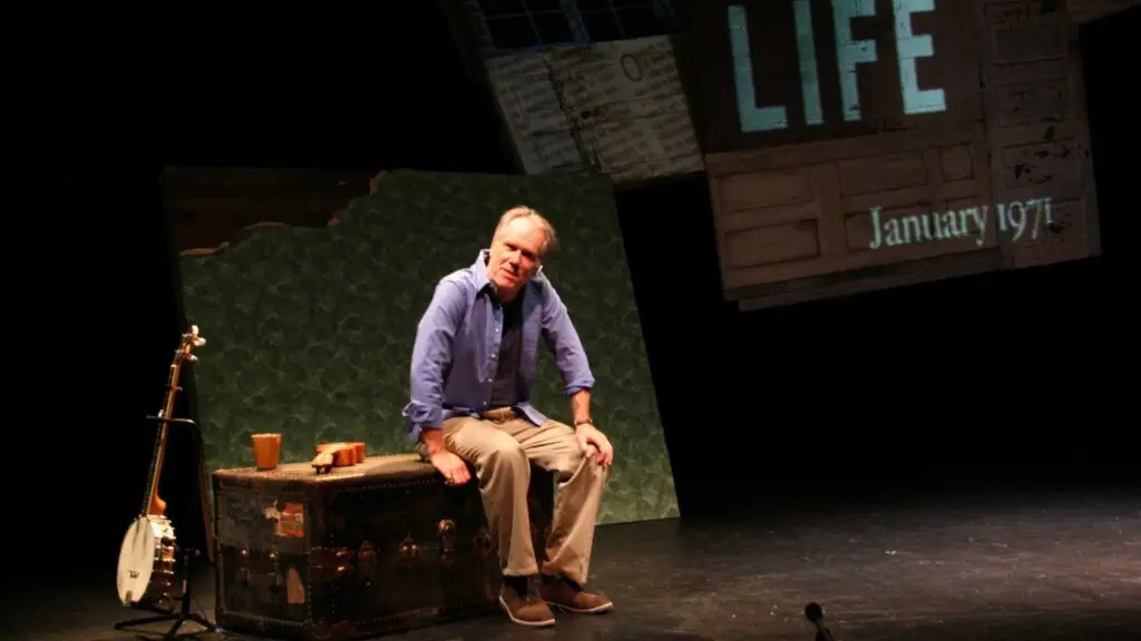 Loudon Wainwright III seated on antique trunk on stage, banjo at his side, during theatrical performance