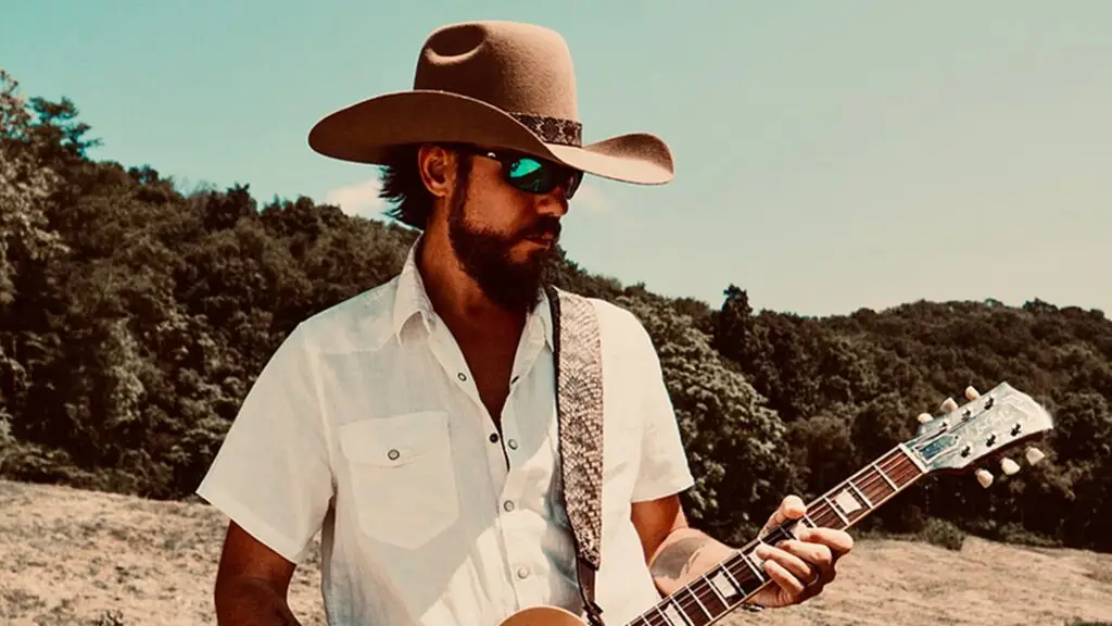 Chris Janson playing guitar outdoors, wearing a cowboy hat and sunglasses, in a rural hillside setting