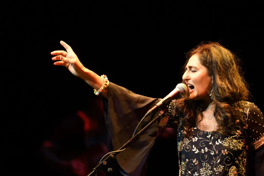 Kiran Ahluwalia singing passionately at microphone, arm raised, in embroidered black outfit