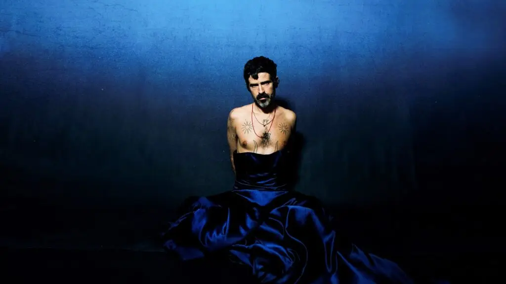 Devendra Banhart in a theatrical studio portrait, draped in a blue satin gown with chest tattoos