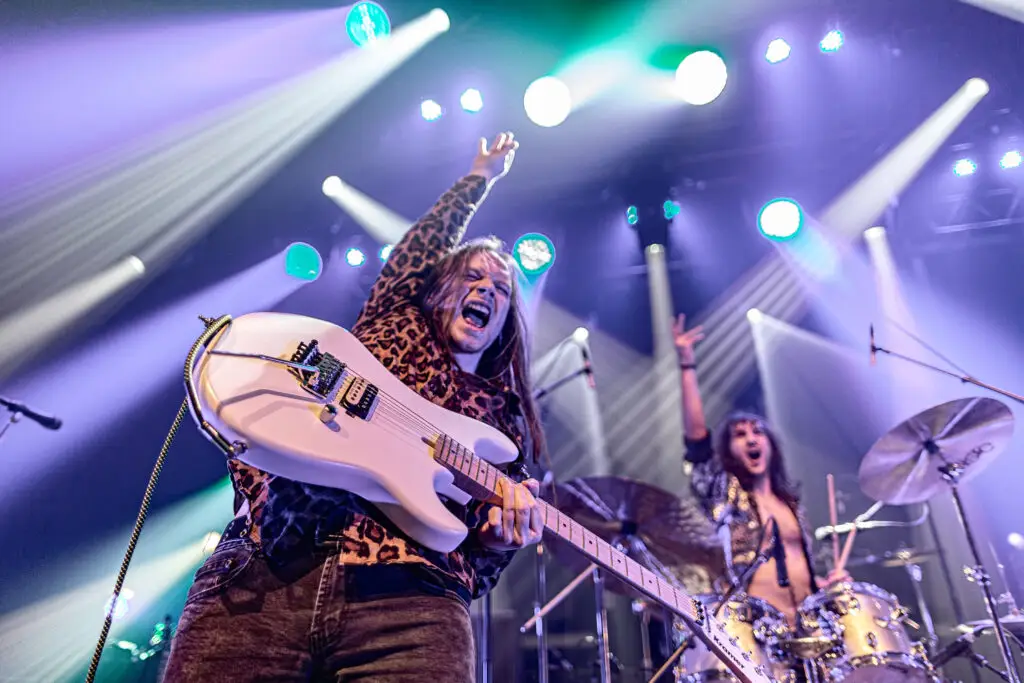 Jacob Deraps performing live with white Kramer guitar, arm raised mid-song, on stage with dramatic lighting