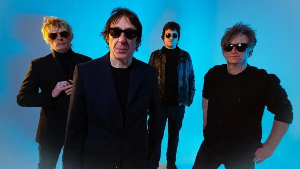 Buzzcocks band press photo, four members in black outfits and sunglasses against blue gradient backdrop
