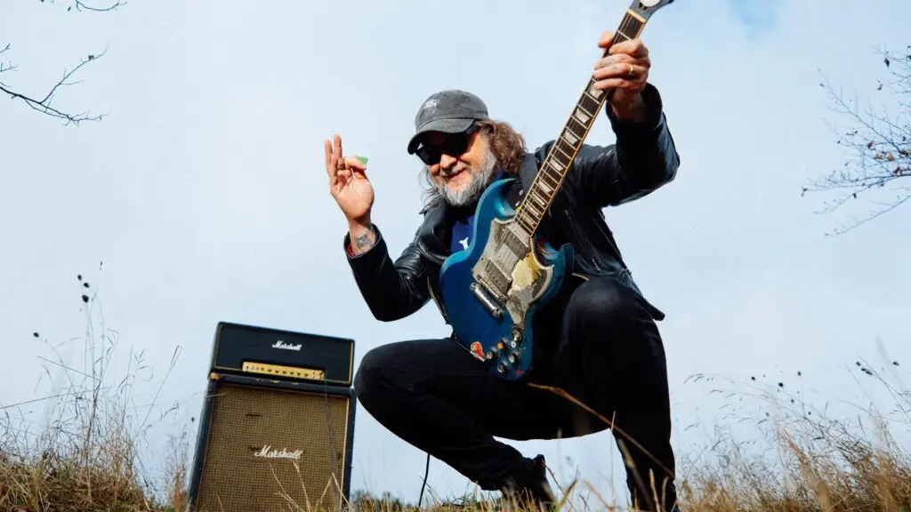 King Tuff crouching outdoors with a blue Gibson SG guitar and Marshall amp stack