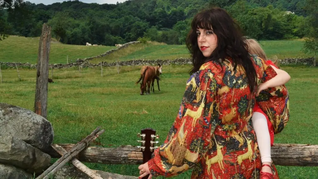 RoseAnn Fino holding acoustic guitar at a Hudson Valley farm fence, wearing colorful tapestry coat