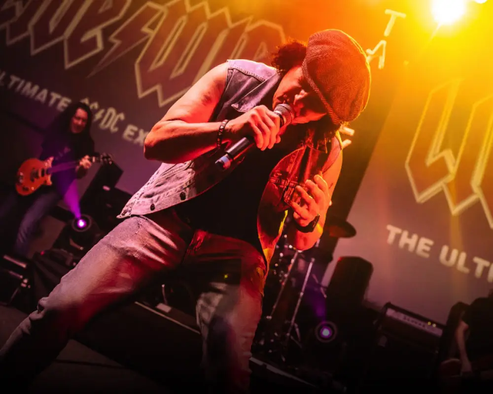 Live Wire vocalist performing live on stage, leaning into mic, with AC/DC tribute backdrop