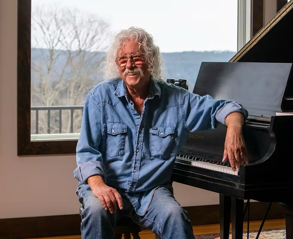 Arlo Guthrie seated at a Baldwin piano, smiling, at his Berkshires home