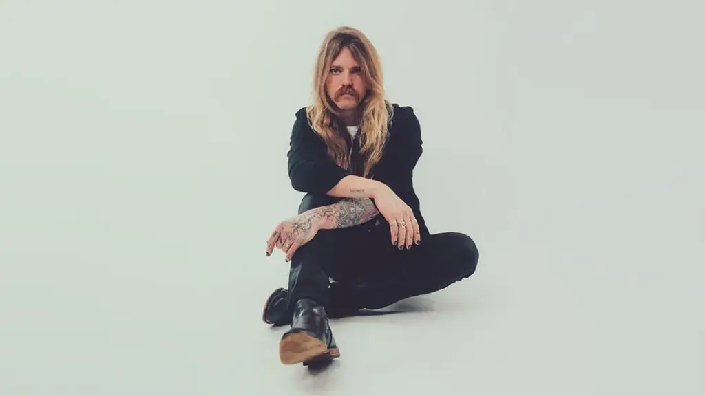 Austin Meade seated in an official press photo, showing long hair and tattooed arms