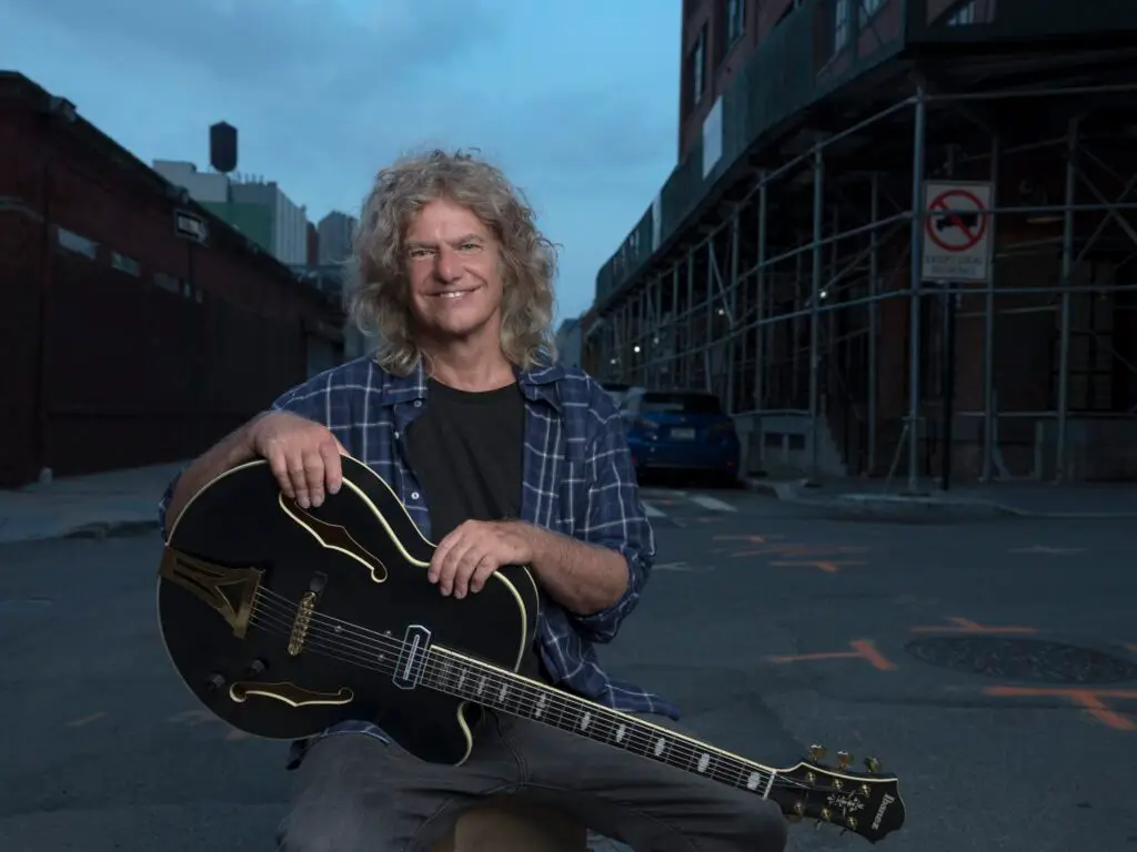 Pat Metheny seated outdoors holding his Ibanez archtop jazz guitar, smiling