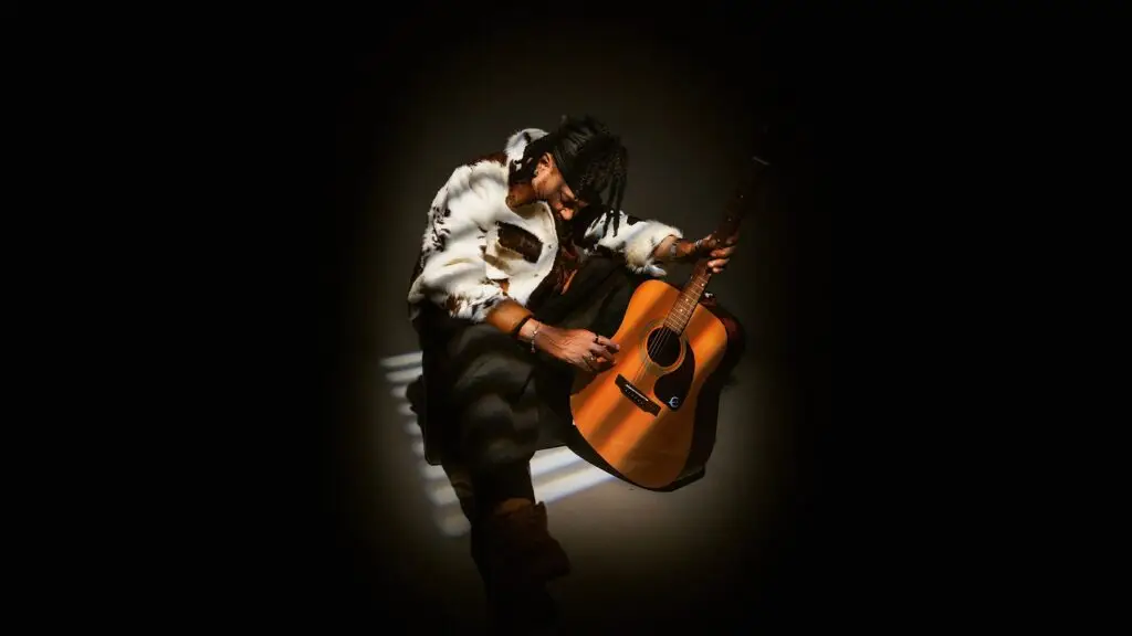 Alan Cavé poses with acoustic guitar in dramatic spotlight, official promotional photo