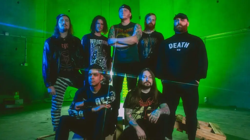 AngelMaker deathcore band promo photo, seven members posed in green-lit industrial space