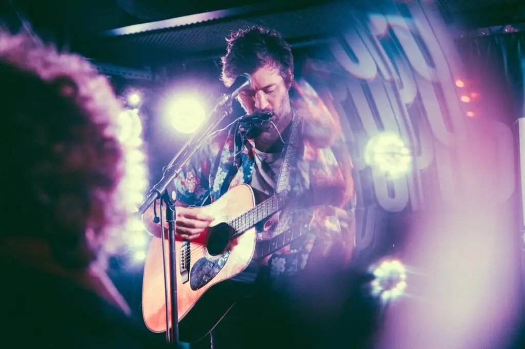 John Craigie performing live with acoustic guitar at a microphone under pink and purple stage lights