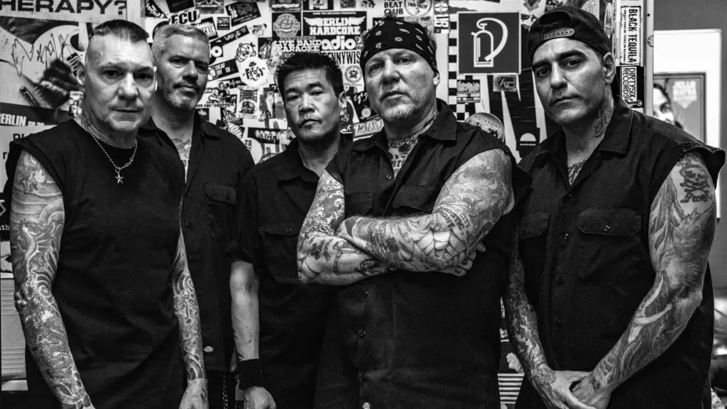 Agnostic Front — five-piece NYC hardcore band posed in front of a sticker-covered wall