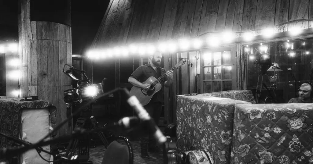 Cris Jacobs playing acoustic guitar in a rustic barn studio with string lights