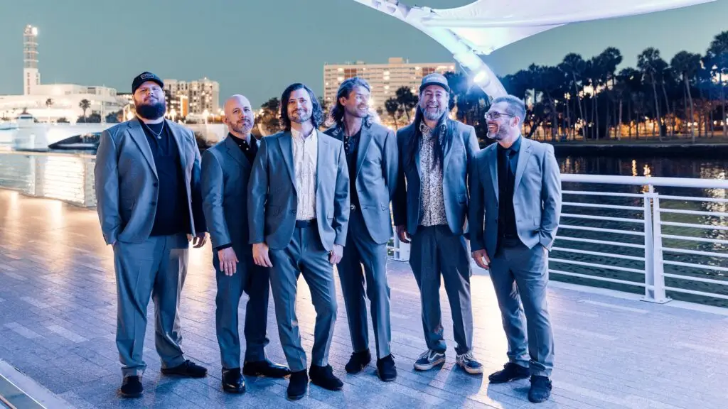 Taking Back Sunday band members pose for their 2026 Louder Now anniversary tour press photo at a waterfront