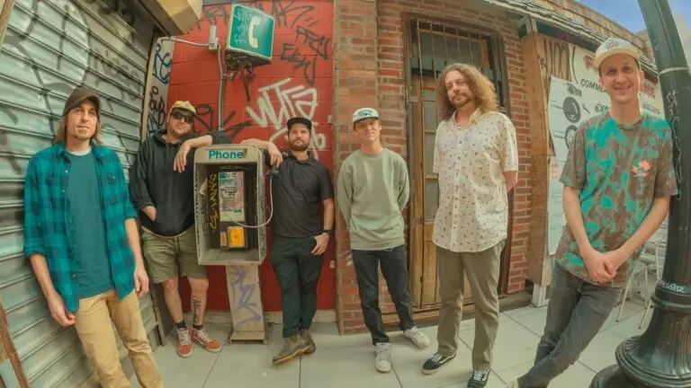 Big Something, six-piece jam band, poses on an urban sidewalk in front of a graffiti-covered brick wall and payphone
