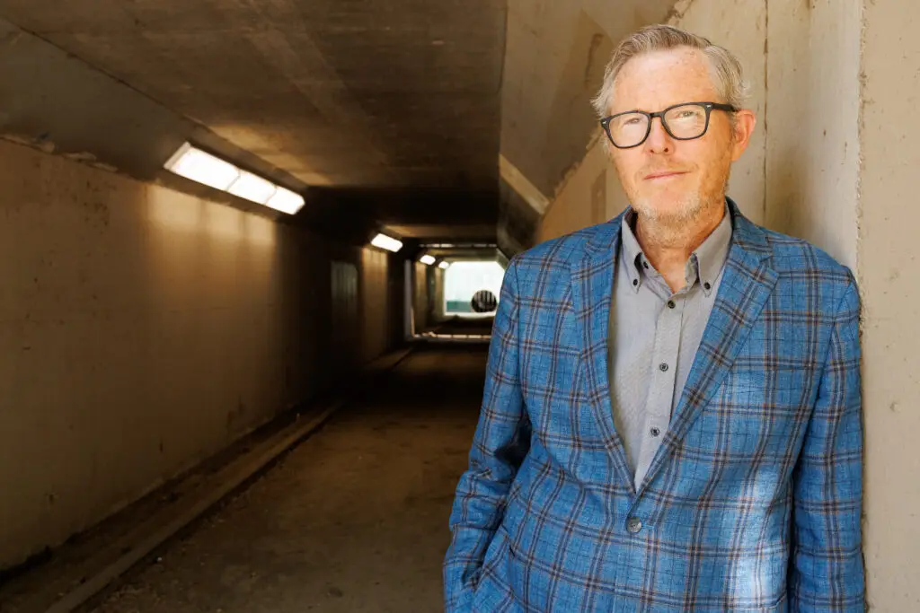 Robbie Fulks in a blue plaid blazer, standing in an urban underpass, official press photo
