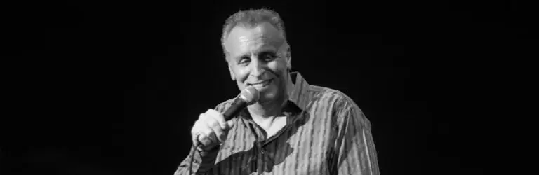 Vic DiBitetto performing stand-up comedy on stage, holding microphone and smiling mid-delivery