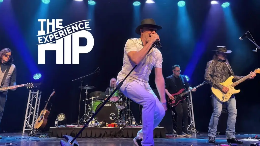 The Hip Experience tribute band performing live, frontman Derek Lathrop leaning into mic with full five-piece band on a blue-lit stage