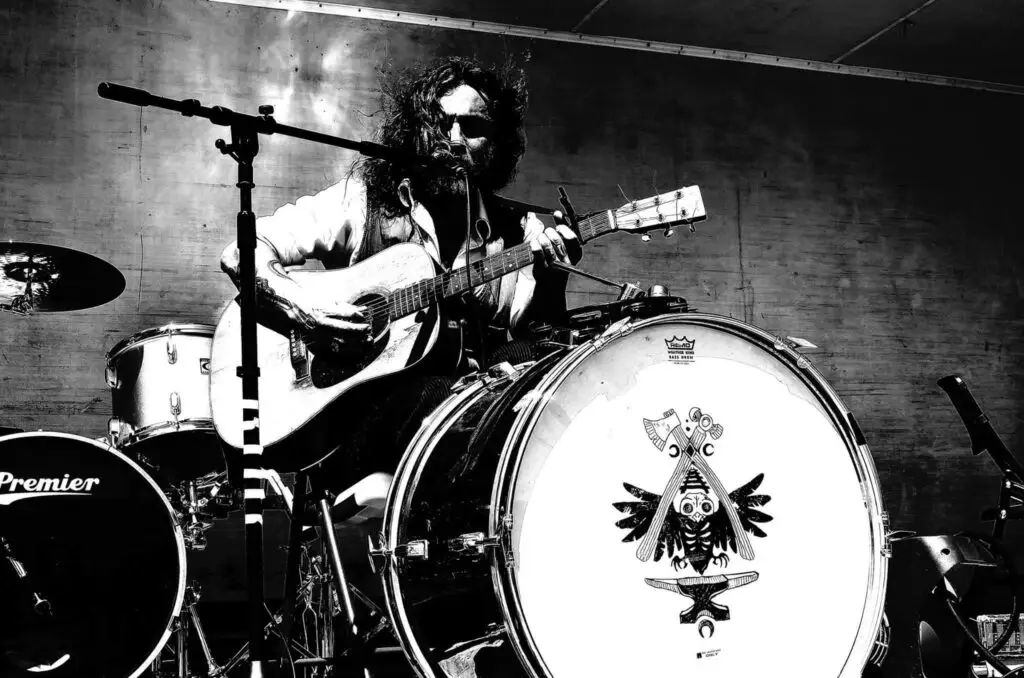 Arthur Buezo performing live as a one-man band, playing acoustic guitar with drum kit, black and white